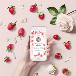 May include: A pink and white gingham background with a strawberry design. The text reads "Please join us for Kylie's Berry First Birthday". The date is November 23rd. The invitation is on a mobile phone.