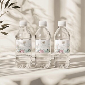 K&ouml;nnte beinhalten: Drei durchsichtige Plastikwasserflaschen mit personalisierten Etiketten f&uuml;r eine Babyparty. Die Etiketten zeigen ein Blumenmuster mit rosa und blauen Blumen und den Text "Amelia's Baby Shower".