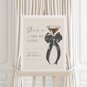 May include: A beige sign with black text and a black bow with a martini glass in the center. The text reads "She's a tiny bit older. Welcome to Victoria's Birthday. Sat Jul 23 20XX".
