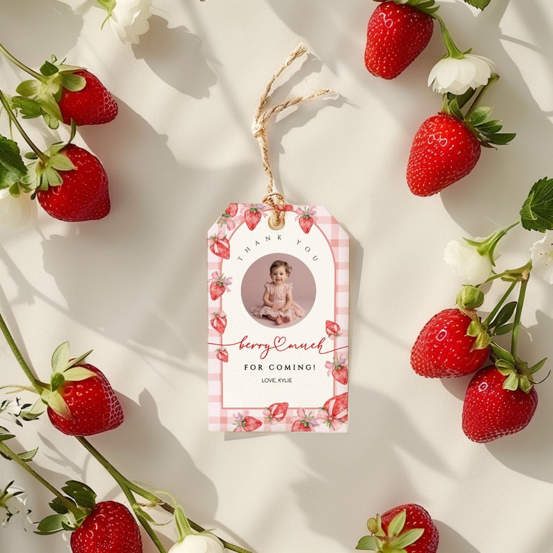 May include: Thank you for coming! A pink and white gingham tag with a photo of a child in a pink dress and a strawberry design. The text on the tag reads "Thank you, Berry much for coming! Love, Kylie".