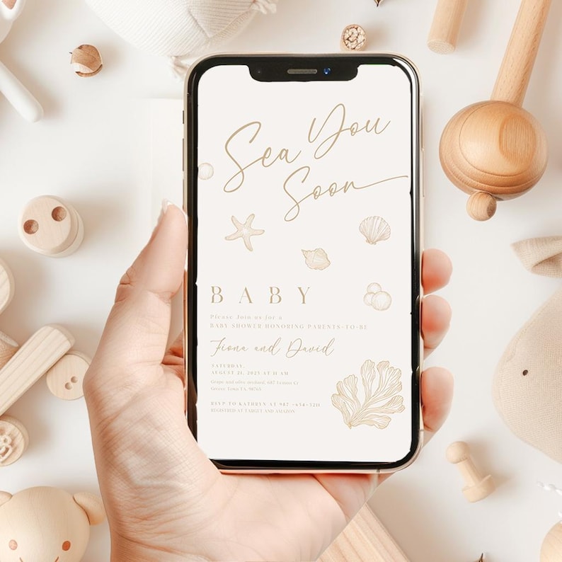 May include: A mobile phone screen shows a baby shower invitation with the words "Sea You Soon" in gold lettering. The design features starfish, seashells, and coral. The phone is held in a hand, with wooden toys around it.
