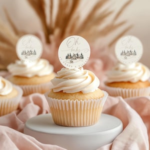 May include: Several cupcakes with white frosting and round toppers. The toppers have the text "Oh baby" and a small illustration of a village scene. The text "IT TAKES A VILLAGE" is also on the toppers. The cupcakes are on a white surface and a pink cloth.