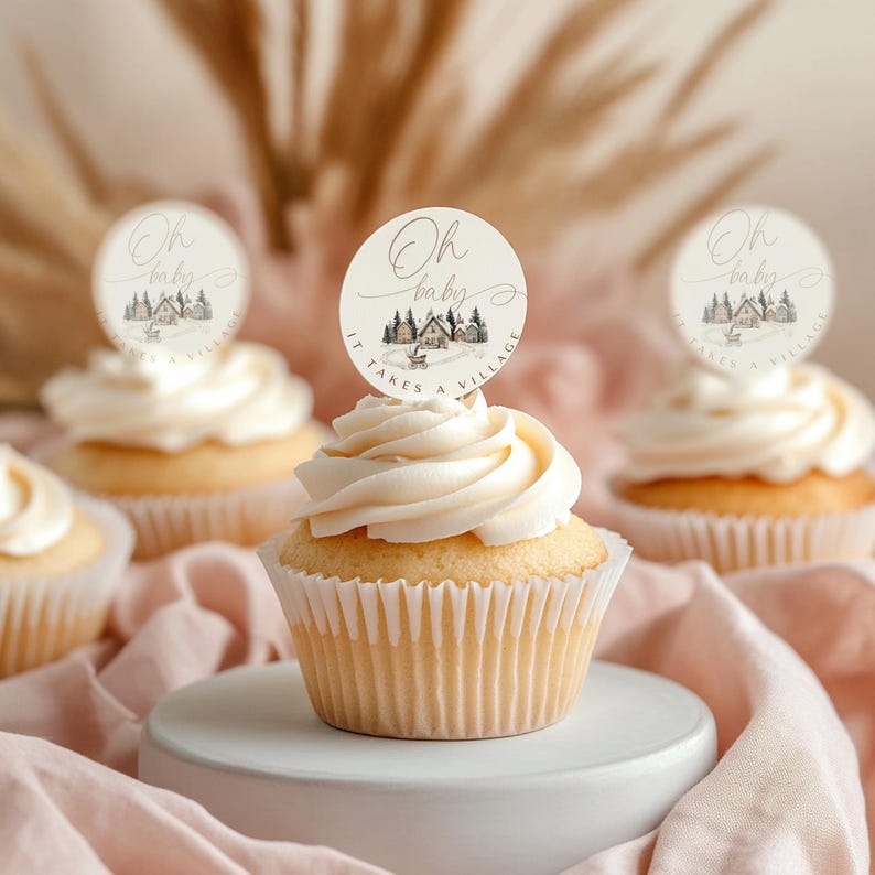 May include: Several cupcakes with white frosting and round toppers. The toppers have the text "Oh baby" and a small illustration of a village scene. The text "IT TAKES A VILLAGE" is also on the toppers. The cupcakes are on a white surface and a pink cloth.