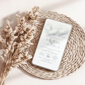 May include: A gender reveal invitation with the text "a little piece of heaven" and "he or she". The invitation is white with a cloud design and is on a woven mat with dried flowers.