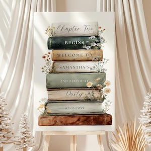 Puede incluir: Un letrero blanco sobre un caballete presenta una pila de libros de acuarela con detalles florales. El texto dice "Chapter Two Begins Welcome to Samantha's 2nd Birthday Party" con una fecha. El letrero está decorado con pequeñas flores.