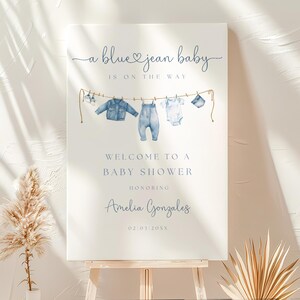 May include: A baby shower sign with the text "a blue jean baby is on the way." The sign features watercolor illustrations of baby clothes hanging on a clothesline. The sign welcomes guests to a baby shower honoring Amelia Gonzales.