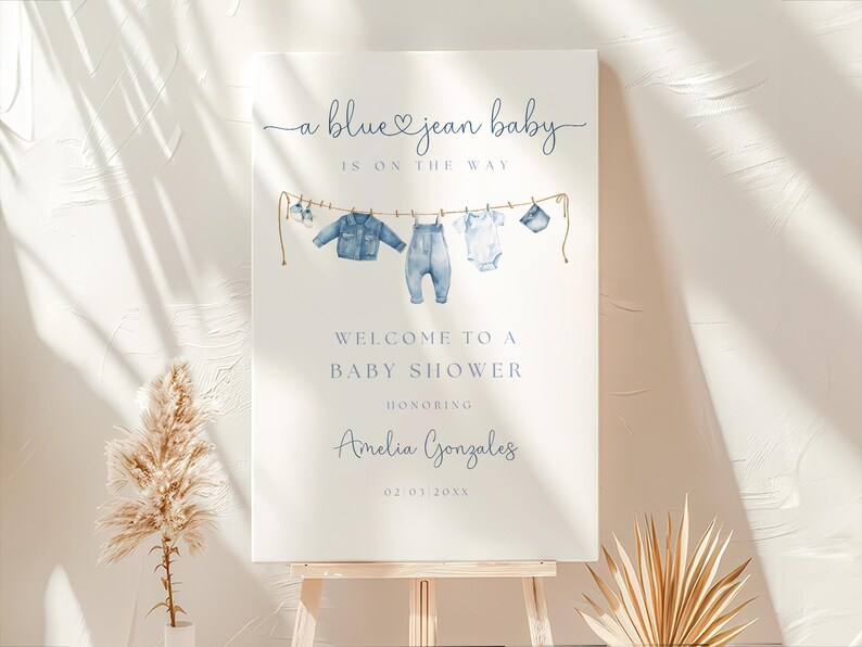 May include: A baby shower sign with the text "a blue jean baby is on the way." The sign features watercolor illustrations of baby clothes hanging on a clothesline. The sign welcomes guests to a baby shower honoring Amelia Gonzales.