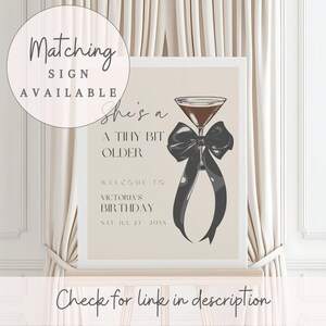 Puede incluir: Un cartel beige con el texto "She's a Tiny Bit Older" y un lazo negro con una copa de martini. El texto "Welcome to Victoria's Birthday Sat Jul 23 20XX" est&aacute; debajo del lazo.