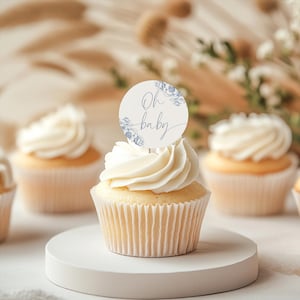 May include: Blue and white cupcake toppers with the text "Oh baby" in a script font. The toppers are on top of cupcakes with white frosting.