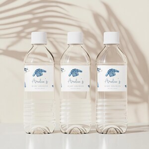 May include: Three clear plastic water bottles with white caps. Each bottle has a label with a blue denim jacket illustration and the text "Amelia's BABY SHOWER 01.01.2023". The bottles are arranged on a white surface.