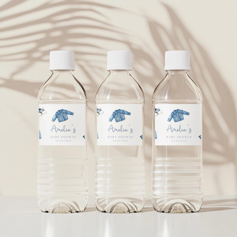 May include: Three clear plastic water bottles with white caps. Each bottle has a label with a blue denim jacket illustration and the text "Amelia's BABY SHOWER 01.01.2023". The bottles are arranged on a white surface.
