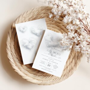 May include: Two white gender reveal invitations with cloud heart designs, resting on a woven tray with white floral accents. The text reads "A little piece of heaven, is on the way, he or she".