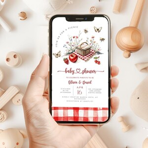 May include: A smartphone displays a baby shower invitation with a picnic theme. The invitation features a picnic basket, red apple, strawberries, and cherries. Text includes "baby shower" and details for Olivia & David's event on April 16th.