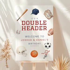 Puede incluir: Un letrero blanco con el texto "IT'S A DOUBLE HEADER" en rojo. El letrero presenta ilustraciones de equipos deportivos, incluyendo un bate de béisbol, una pelota de béisbol, fútbol americano, baloncesto, fútbol, un casco, un guante, una gorra y un silbato. El letrero da la bienvenida al cumpleaños de Joshua y Daniel.
