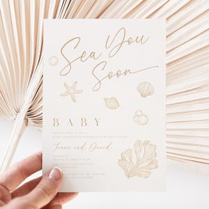 May include: A cream-colored baby shower invitation with gold script that reads "Sea You Soon." The invitation features illustrations of seashells, a starfish, and coral. The names "Fiona and David" are also included. The invitation is held by a hand.