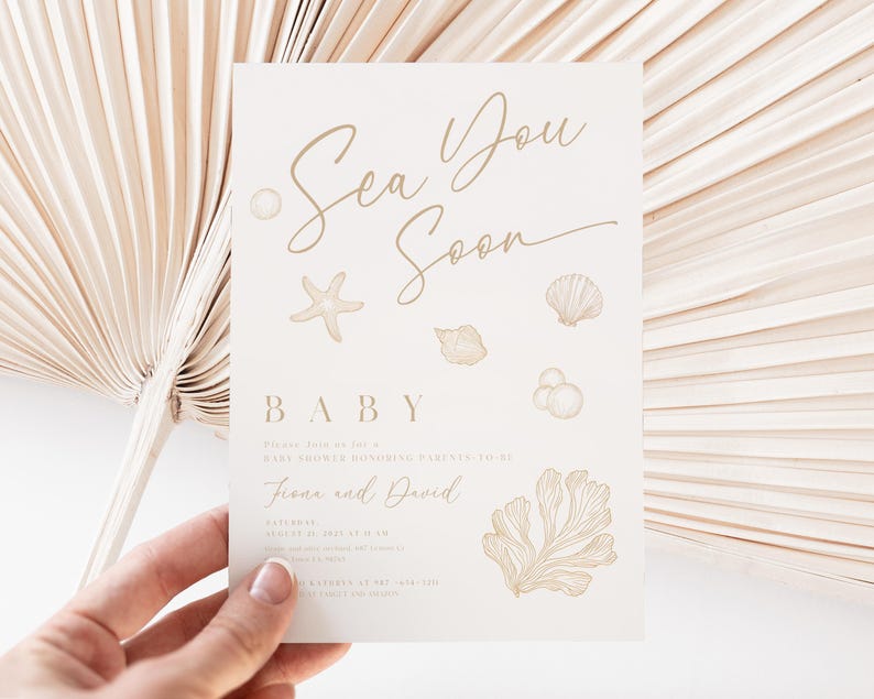 May include: A cream-coloured baby shower invitation with gold script that reads "Sea You Soon." The invitation features illustrations of seashells, a starfish, and coral. The names "Fiona and David" are also included. The invitation is held by a hand.