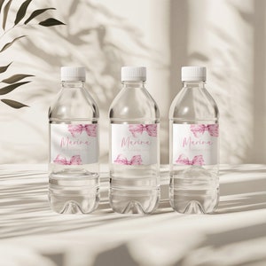 May include: Three clear plastic water bottles with pink and white labels. The labels have a watercolor design of a bow and the text "Marina 1st Birthday Party".