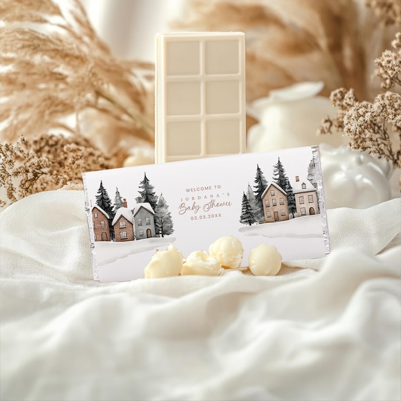 May include: A white chocolate bar and a wrapped chocolate bar with a winter scene and the text "Welcome to Jordana's Baby Shower". The wrapped bar features illustrations of houses and trees. Several white chocolate pieces are in front of the wrapped bar.