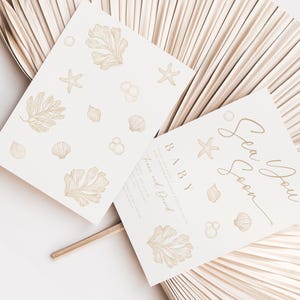 May include: Two cream-coloured baby shower invitations with a sea theme. One card features the text "Sea You Soon BABY" in gold script, and the other has sea-themed illustrations. The cards are decorated with starfish, shells, and seaweed.