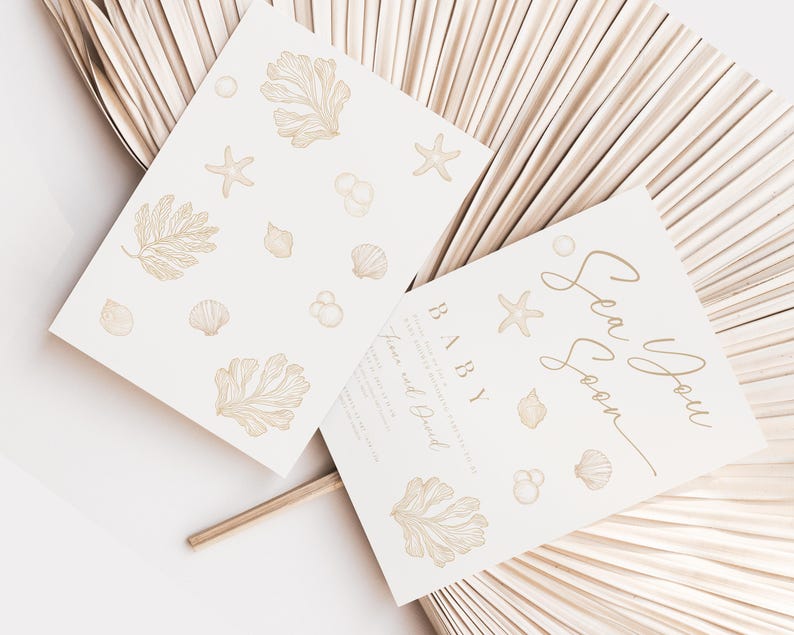 May include: Two cream-coloured baby shower invitations with a sea theme. One card features the text "Sea You Soon BABY" in gold script, and the other has sea-themed illustrations. The cards are decorated with starfish, shells, and seaweed.