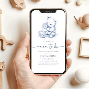 Peut inclure: Une invitation de f&ecirc;te pr&eacute;natale bleue et blanche avec une illustration de dessin anim&eacute; d'un ours en peluche versant du th&eacute;. Le texte indique "Tea with the mom to be. Please join us for a baby shower honoring Fiona Johns. Sat Jul 23 11 am. Grape & Olive Backyard Bar. 33 Maria Rd. Fazenda, New Zealand. RSVP to Katrina on 021 029 786 35."