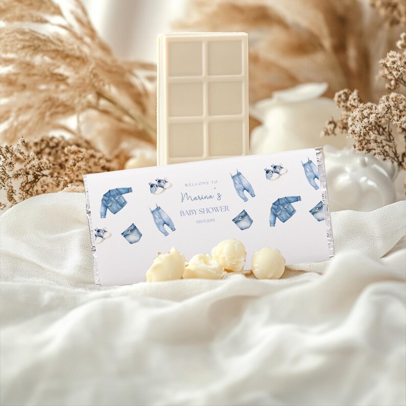May include: A white chocolate bar and pieces are displayed with a baby shower-themed wrapper. The wrapper features blue denim-themed illustrations and text that reads "Welcome to Marina's Baby Shower".