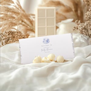 May include: White chocolate bar wrapped in a silver foil wrapper with a white card attached. The card has a blue and white teacup illustration and the text "Welcome to Jordana's Baby Shower 02.25.2023".
