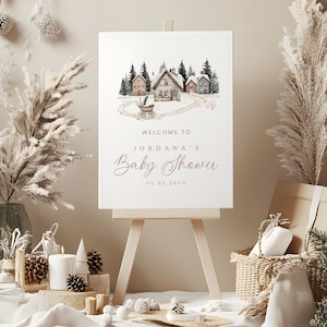 May include: A baby shower sign on a wooden easel. The sign features a watercolor illustration of a snowy village scene with a baby carriage. The text reads "Welcome to Jordana's Baby Shower" and the date.