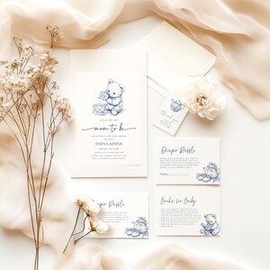 Peut inclure: Invitation de f&ecirc;te pr&eacute;natale bleu et blanc avec un ours en peluche buvant du th&eacute;. L'invitation indique "Tea with the Mom to Be". Il y a aussi des cartes pour une tombola de couches et des livres pour b&eacute;b&eacute;.