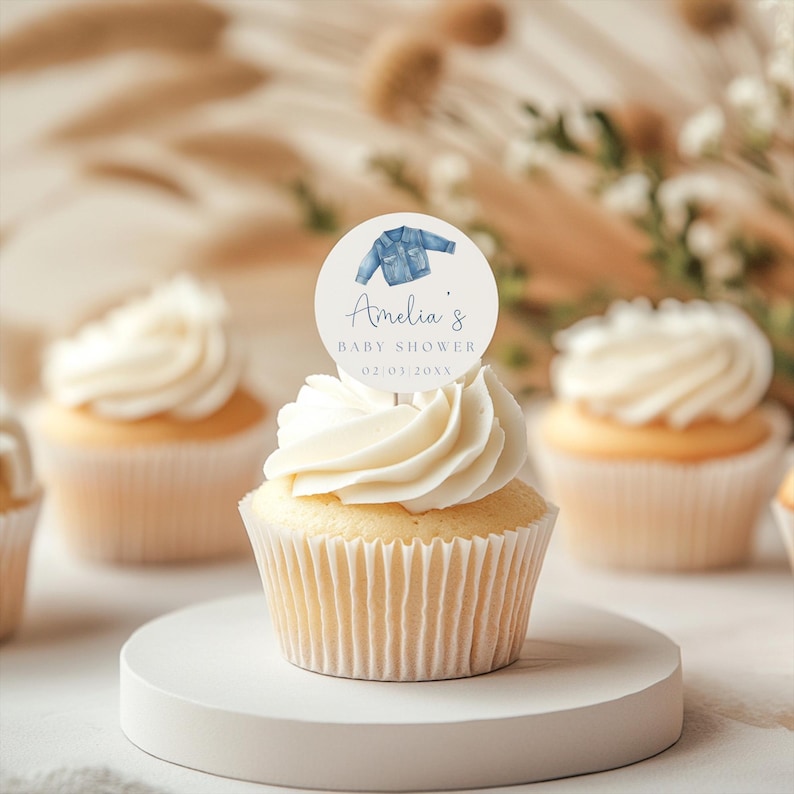 May include: A cupcake with white frosting and a round topper. The topper features a watercolor illustration of a denim jacket, the name "Amelia's", and the words "Baby Shower" and a date. Other cupcakes are in the background.
