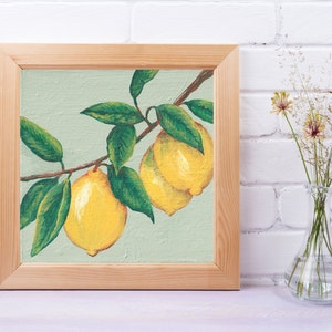 May include: A square painting in a light-colored wooden frame depicting two bright yellow lemons hanging from a brown branch with several dark green leaves against a pale sage green background, showcasing visible brushstrokes and texture.