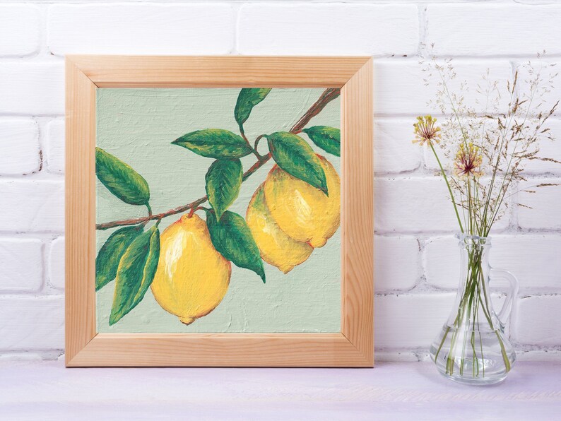 May include: A square painting in a light-colored wooden frame depicting two bright yellow lemons hanging from a brown branch with several dark green leaves against a pale sage green background, showcasing visible brushstrokes and texture.