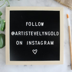 May include: A square felt letter board with a light wood frame and a black background. The white plastic letters spell out FOLLOW @ARTISTVELYNGOLD ON INSTAGRAM with a small white heart symbol centered at the bottom of the board.