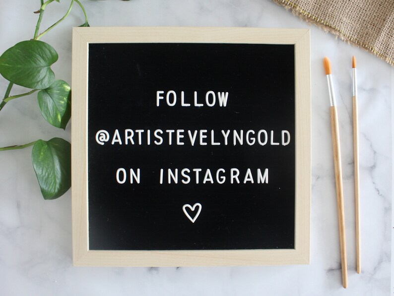 May include: A square felt letter board with a light wood frame and a black background. The white plastic letters spell out FOLLOW @ARTISTVELYNGOLD ON INSTAGRAM with a small white heart symbol centered at the bottom of the board.