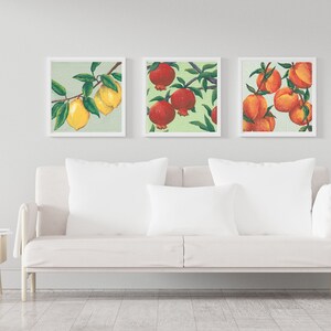 May include: Three square framed wall art prints featuring botanical illustrations. The left print shows yellow lemons on a branch, the center print displays red pomegranates, and the right print depicts orange peaches, all set against a light green background.