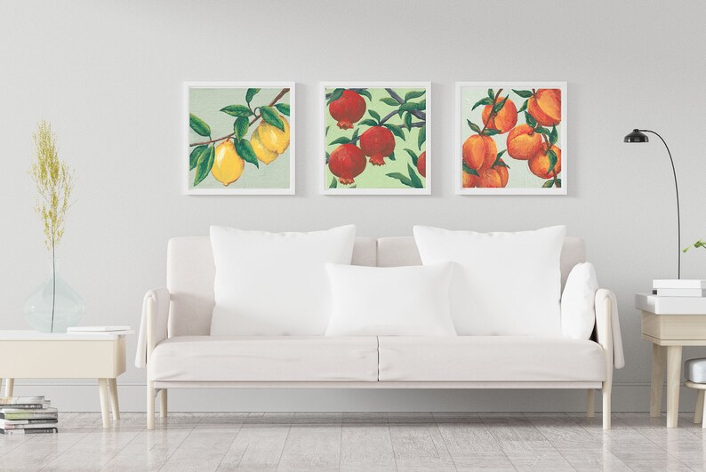 May include: Three square framed wall art prints featuring botanical illustrations. The left print shows yellow lemons on a branch, the center print displays red pomegranates, and the right print depicts orange peaches, all set against a light green background.