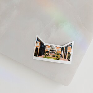 May include: A sticker featuring a cartoon illustration of a brick building with a courtyard. The building has a red roof and white trim. The courtyard is green and has trees and bushes.