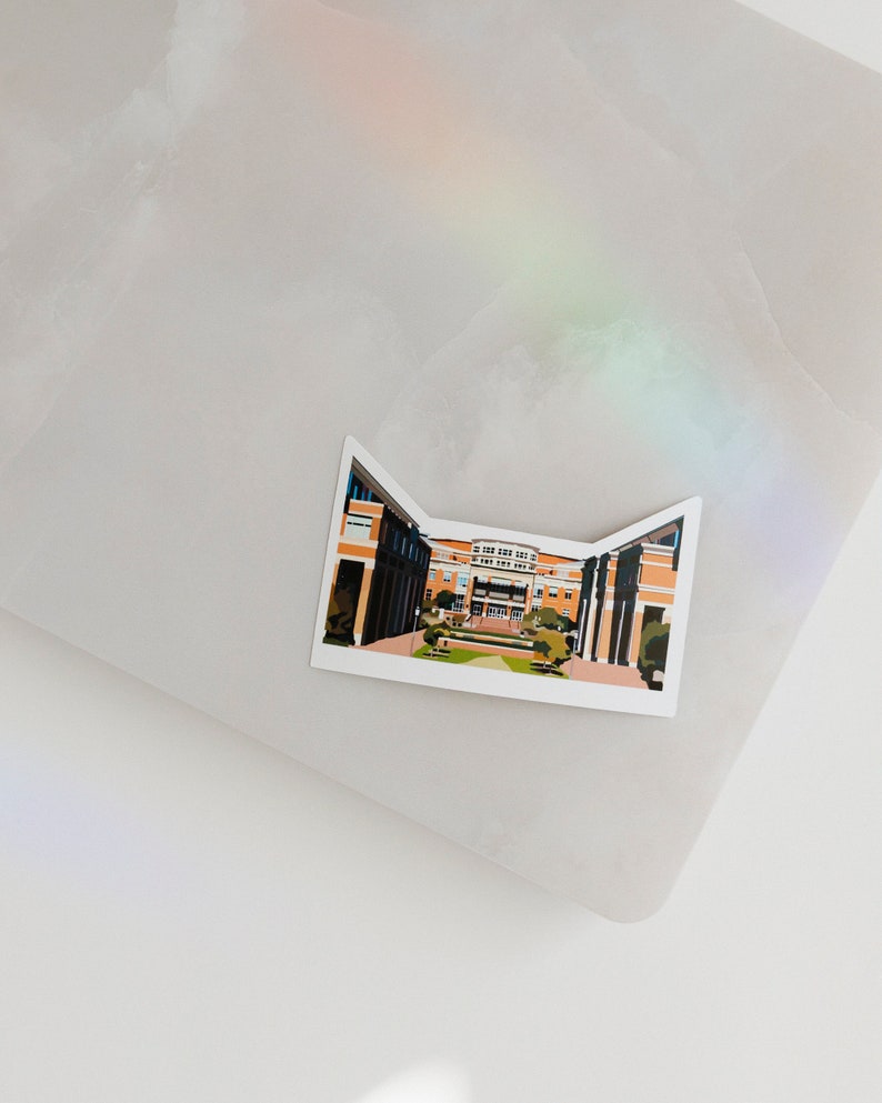 May include: A sticker featuring a cartoon illustration of a brick building with a courtyard. The building has a red roof and white trim. The courtyard is green and has trees and bushes.