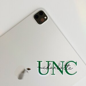 May include: A white laptop with a green UNC Charlotte sticker on the back. The laptop has a silver Apple logo on the back.