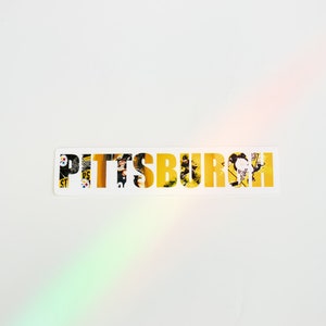 May include: A white sticker with the word "PITTSBURGH" in yellow letters. The letters are made up of images of Pittsburgh landmarks and scenes.