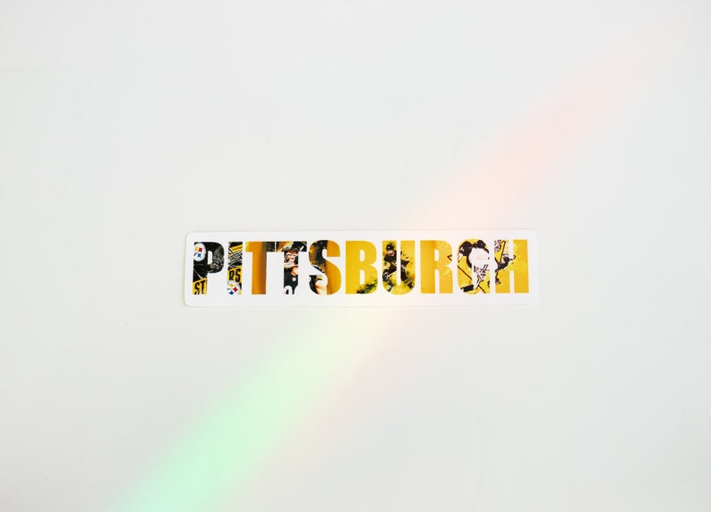 May include: A white sticker with the word "PITTSBURGH" in yellow letters. The letters are made up of images of Pittsburgh landmarks and scenes.