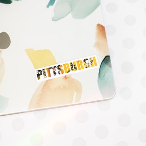 May include: A white sticker with the word "PITTSBURGH" in yellow letters. The letters are made up of smaller images, creating a collage effect. The sticker is on a white surface with a light blue and yellow watercolor design.