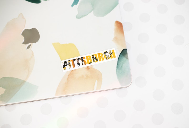 May include: A white sticker with the word "PITTSBURGH" in yellow letters. The letters are made up of smaller images, creating a collage effect. The sticker is on a white surface with a light blue and yellow watercolor design.