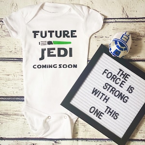 May include: White baby onesie with the text "Future Jedi Coming Soon" and a green lightsaber graphic. A black letter board with the text "The Force is Strong With This One" is next to the onesie.