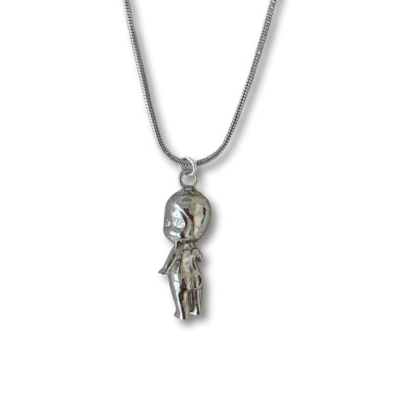 May include: A silver necklace with a pendant in the shape of a stylized figure. The pendant is attached to a fine chain. The figure has a rounded head and a slender body. The necklace is set against a white backdrop.