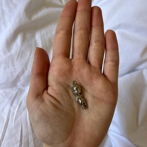 K&ouml;nnte beinhalten: Ein kleiner, silberfarbener Anh&auml;nger in Form einer stilisierten menschlichen Figur. Der Anh&auml;nger hat oben eine Schlaufe zur Befestigung und wird in der Handfl&auml;che vor wei&szlig;em Hintergrund gehalten.