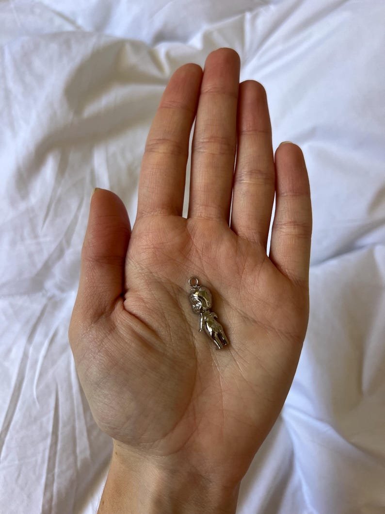 K&ouml;nnte beinhalten: Ein kleiner, silberfarbener Anh&auml;nger in Form einer stilisierten menschlichen Figur. Der Anh&auml;nger hat oben eine Schlaufe zur Befestigung und wird in der Handfl&auml;che vor wei&szlig;em Hintergrund gehalten.
