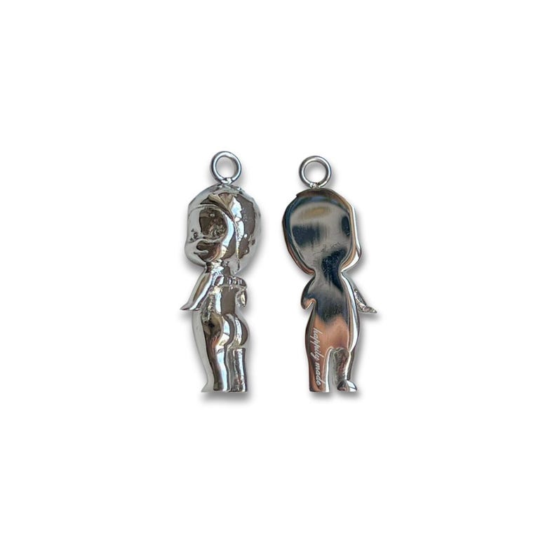 K&ouml;nnte beinhalten: Zwei silberfarbene Anh&auml;nger in Form einer stilisierten Figur, einer mit Vorder- und einer mit R&uuml;ckseite. Jeder Anh&auml;nger hat oben eine kleine Schlaufe zum Aufh&auml;ngen. Der vordere Anh&auml;nger hat detaillierte Merkmale, w&auml;hrend der hintere Anh&auml;nger glatt ist.