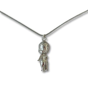 May include: A silver pendant necklace with a doll-shaped charm. The charm has a rounded head, small arms, and a simple body. The necklace chain is a thin, silver snake chain. The background is white.
