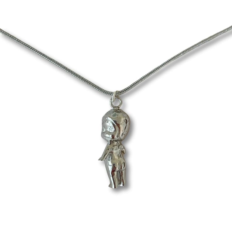 May include: A silver pendant necklace with a doll-shaped charm. The charm has a rounded head, small arms, and a simple body. The necklace chain is a thin, silver snake chain. The background is white.